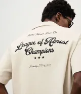 Camisa de corte boxy en color beige, con cuello inglés y mangas cortas. Presenta un diseño con lettering bordado en la espalda con la frase 'League of Almost Champions' y detalles gráficos en negro.