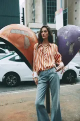 Camisa de manga larga con estampado de rayas verticales en color naranja y crema. Presenta cuello solapa, cierre frontal con botones dorados y puños anchos en color crema con detalle de lazos ajustables.