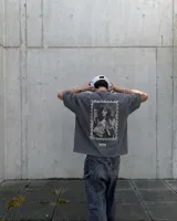 Remera oversize gris oscuro con corte holgado, mangas cortas y estampa trasera en blanco y negro con forma de estampilla que muestra a una persona con un gato y el texto 'Kaboa Industries'.