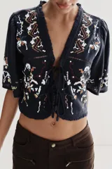 Top corto de color negro con escote en pico y mangas cortas anchas. Presenta bordados coloridos en estilo geométrico y floral, detalle de entredoses y cierre frontal mediante lazos con abalorios.