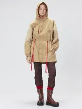 Trench tipo piloto color beige con capucha, cierre frontal con botones y cordones ajustables en color rojo en cintura y capucha. Presenta puños elásticos, bolsillos laterales amplios y un bolsillo invisible en el pecho con logo bordado de la marca.