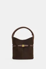 Bolso tipo saca de tamaño mini, confeccionado en gamuza color marrón oscuro con detalles en cuero marrón. Presenta un asa de mano fija, una tira decorativa horizontal con hebilla metálica dorada en el frente y costuras verticales visibles.