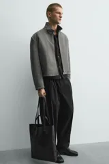 Pantalón negro de corte relaxed fit y estilo wide leg, con pliegues frontales dobles, bolsillos delanteros y detalle de bolsillos traseros de vivo. Cierre frontal cruzado con cremallera y gancho metálico oculto.