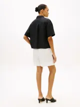 Camisa de manga corta de corte amplio confeccionada en puro lino, de color negro, con cuello inglés y un bolsillo de parche en el pecho que incluye un sutil logo bordado. Presenta hombros ligeramente caídos y cierre frontal de botones.