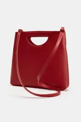 Bolso de mano de piel color rojo, con diseño de cuerpo asimétrico y asa de mano integrada con apertura lateral. Incluye correa bandolera extraíble y bolsillo interior.
