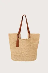 Bolsa tote de fibra de rafia tejida color beige, con asas dobles fijas de cuero sintético marrón y etiqueta colgante con logotipo grabado.