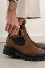 Bota tipo Chelsea de cuero marrón con suela track negra de goma. Presenta paneles elásticos laterales y una costura reforzada en la puntera. Incluye una correa de cuero desmontable con hebilla metálica.