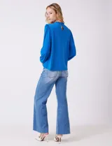 Blusa azul de gasa con pliegues en la parte delantera y detalles de encaje calado. Cuello alto fruncido y mangas largas con puños elastizados. Espalda lisa con abertura tipo gota y botón.