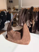 Cartera tipo hobo de diseño relajado, confeccionada en material con textura simil gamuza color chocolate. Presenta una silueta suave, cierre superior y asa corta combinada en simil cuero al tono.