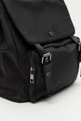 Mochila urbana de nylon color beige, con solapa superior, bolsillo frontal con cierre y correas decorativas con hebillas metálicas. Incluye manija superior y correas ajustables.