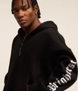 Saco tipo hoodie con cierre frontal de cremallera, color negro, corte boxy y capucha. Presenta estampa de letras en blanco en ambas mangas y un colgante de cruz metálico en el tirador de la cremallera principal.