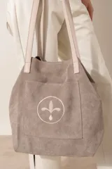 Bolso tipo tote de gamuza color chocolate, con bolsillo frontal externo que presenta un diseño circular bordado con una flor estilizada. Cuenta con correas largas de cuero en tono crema.