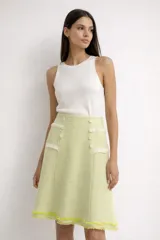 Falda midi de tweed color verde limón y blanco, de corte acampanado con cintura alta. Presenta detalles de bolsillos simulados decorados con solapas con flecos y botones revestidos, y terminaciones deshilachadas en el borde inferior.