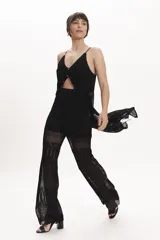 Pantalón de tejido de punto negro, corte ancho y tiro alto, con detalle de encaje calado en la parte inferior. Se lleva debajo de un vestido largo de encaje negro con tirantes finos y botones frontales.