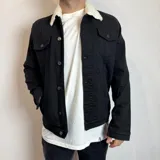 Chaqueta de jean negra con cuello de corderito blanco, cierre frontal con botones metálicos y bolsillos frontales con solapa.