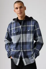 Camisa de franela a cuadros en tonos azul, gris y blanco, con capucha negra y cordones ajustables. Presenta dos bolsillos frontales con solapa y botones, y cierre de botones en el frente.