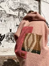 Campera tipo abrigo de textura borreguito en color rosa, con diseño de collage en la espalda que incorpora un parche de pana rojiza y un panel central con estampado geométrico étnico. Presenta capucha integrada y un detalle de etiqueta de cuero sintético en la parte posterior.