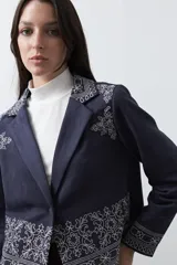 Blazer de gamuza azul marino con bordados florales a contratono en solapas, puños y ruedo. Presenta corte recto, solapas clásicas y cierre frontal con botón forrado.
