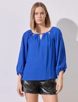 Blusa de gasa color azul, con cuello redondo y aberturas frontales en V. Presenta mangas raglán con apliques de puntilla a lo largo y puños fruncidos.