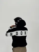 Canguro con capucha de corte boxy, color negro con una franja horizontal blanca que recorre la espalda y las mangas, presentando la palabra 'BROOKLYN' estampada en negro sobre la franja. La capucha también incluye detalles estampados.