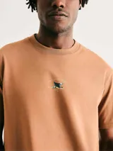 Camiseta de cuello redondo y manga corta en color marrón claro, con un pequeño estampado central en el pecho que incluye un diseño gráfico y texto.
