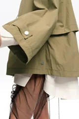 Gabardina corta en tono verde militar, de corte línea A en tejido técnico. Presenta cierre frontal con cremallera, cuello alto con capucha ajustable mediante cordón, mangas largas acampanadas con puños ajustables, bolsillos delanteros tipo parche y aberturas laterales con botones a presión.