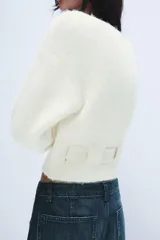 Jersey de punto con escote barco y manga larga. Presenta un diseño corto con detalle de cinturón integrado en el mismo tejido y bolsillos decorativos en la parte frontal.