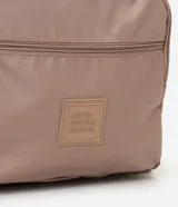 Mochila femenina color beige con textura acolchada y bolsillo frontal con cierre. Incluye asas largas ajustables, correa de mano y llavero de pompón.