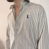 Camisa de manga larga color beige claro, con cuello clásico, cierre frontal con botones y logo bordado en el pecho. Corte regular.