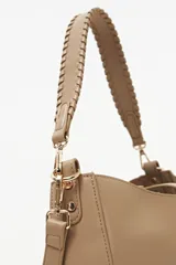 Cartera tipo hobo color beige, de diseño estructurado con cierre superior y una tira vertical con detalle de dije metálico circular en la parte frontal. Cuenta con asa corta trenzada y herrajes dorados.