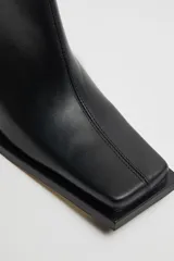 Botas negras de cuero brillante a media pantorrilla con taco de cuero en cascada de 5cm y punta cuadrada alargada. Panel elástico en el lateral trasero de la pantorrilla y lengüeta.