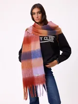 Bufanda de tejido suave con diseño de cuadros grandes en tonos naranja, rojo, violeta y lila. Presenta flecos en los extremos y una etiqueta pequeña con el logo de la marca Rusty.