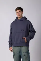 Buzo canguro oversize de frisa de algodón premium en color azul grisáceo. Presenta un diseño con recortes frontales, bolsillo tipo canguro y un pequeño logo circular estampado en el pecho. Cuenta con puños y ruedo acanalados.