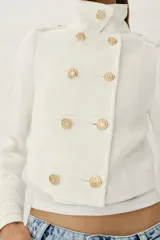 Chaqueta corta blanca de estilo militar, con cuello subido, manga larga y trabillas desflecadas en los hombros. Cuenta con cierre frontal cruzado decorado con botones metálicos dorados.
