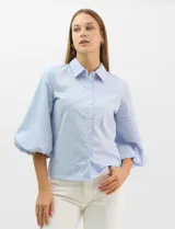 Camisa de algodón con patrón de rayas verticales, cuello camisero clásico y cierre frontal con botones. Presenta detalles metálicos aplicados en el frente y mangas, con mangas 3/4 abullonadas y ruedo recto.