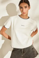 Remera marrón de manga corta y cuello redondo, con detalles de costuras decorativas en contraste color crudo en el cuello, los puños y el dobladillo inferior. Cuenta con el nombre de la marca impreso en el pecho.