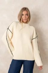 Buzo de punto acanalado color beige, con cuello alto, corte oversize y mangas largas. Presenta costuras decorativas en color negro que recorren los hombros y las mangas.