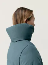 Campera larga tipo puffer de color beige, con acabado mate, cuello alto envolvente y cierre frontal con cremallera. Presenta diseño de bandas acolchadas termoselladas, bolsillos laterales con cierre oculto y relleno vegano.