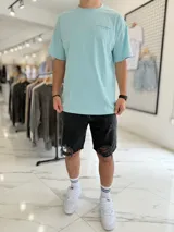 Remera celeste oversize con estampa en la espalda de un paisaje marino con palmeras y olas dentro de un círculo, acompañada de texto.