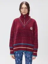 Pullover de textura tipo terciopelo con cuello alto y cierre frontal parcial. Presenta un diseño a rayas horizontales en tonos grises y negros, con detalles en rojo y blanco en el cuello, puños y ruedo. Incluye un aplique bordado con la letra M en el pecho y puños acanalados.