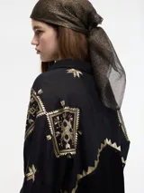 Camisa negra de manga larga con cuello camisero y bordados geométricos en hilo dorado. Presenta un borde en zigzag dorado en la abertura frontal y un largo irregular en la espalda.