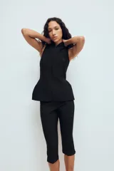 Chaleco entallado de color negro con cuello alto y cierre frontal con cremallera oculta. Presenta un diseño sin mangas con costuras verticales que definen la silueta y un bajo con ligero volumen acampanado.