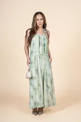 Vestido largo de corte holgado con escote halter y lazo ajustable en el cuello. Presenta un estampado tipo tie-dye en tonos verdes y crema, con un cinturón fino del mismo tejido para ajustar la cintura y bolsillos laterales.