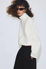 Chaqueta de punto color crudo con cuello alto, cierre frontal de cremallera metálica, mangas largas con puños elásticos y bajo ajustable con cordones.