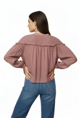 Camisa azul de corte holgado con cuello a la base, cierre frontal con botones y mangas largas abullonadas. Presenta detalles de plisado en los hombros y un delicado volado horizontal que recorre el pecho y la espalda.