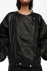 Chaqueta de corte globo cruzado confeccionada en piel ovina de color negro. Presenta cierre frontal con botones a presión, cuello redondeado, mangas largas tipo murciélago, bolsillos con cremallera y fuelles en la espalda que aportan volumen a la silueta.