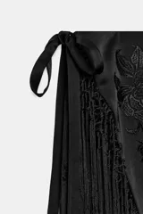 Falda corta de tiro medio, color negro, con diseño cruzado estilo pareo ajustable con lazada lateral. Cuenta con bordados tonales de motivos florales y bajo acabado con flecos largos.