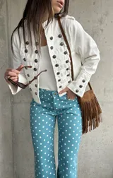 Chaqueta corta de estilo militar en color blanco, con múltiples filas de botones metálicos grabados y detalles de cordones en el frente. Cuenta con cuello mao y mangas largas con botones en los puños.