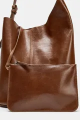 Bolso tipo saca de diseño asimétrico confeccionado en piel de color marrón. Cuenta con un asa de hombro ajustable y cierre mediante imán. Incluye una cartera con bolsillo extraíble en su interior.
