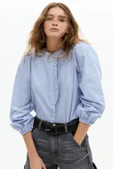 Camisa de algodón a rayas verticales celestes y blancas, con cuello mao, mangas largas abullonadas con puños elásticos y detalle de volado en la cartera de botones frontal.